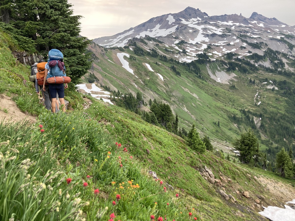 Ascending Goat Ridge A group of backpackers climbing a narrow path up an alpine ridge toward a cluster of mountains dotted with patches of snow