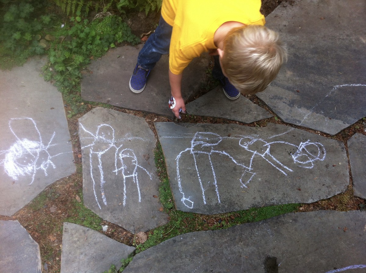 Overhead view of a child and his chalk drawing on basalt flagstones. Picture is 5 human figures and a small bundle, representing the artist’s family