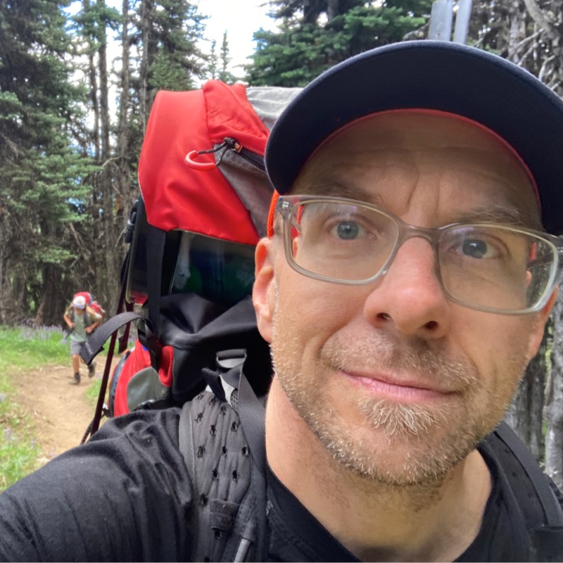 A white man with glasses and five days’ stubble, wearing a baseball cap and large backpack, hiking through a forest. In the background, out of focus, is a young person wearing a backpack.